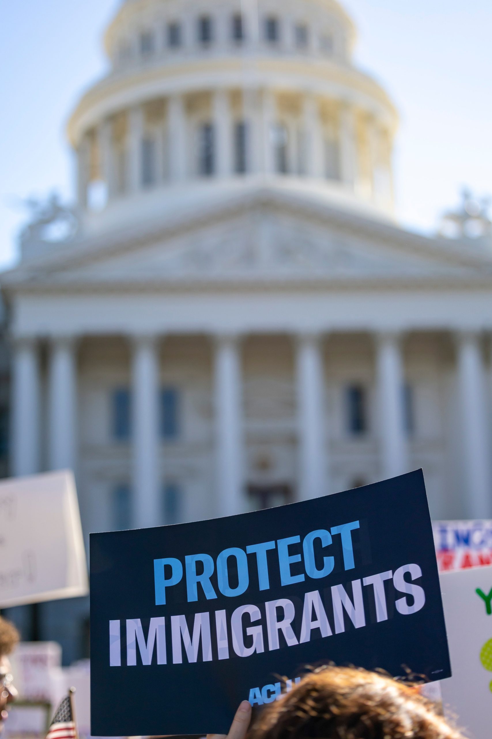 A protest sign reading Protect Immigrants held up at a rally outside a government building.